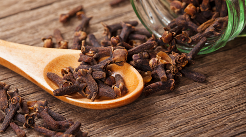 Dried cloves in a wooden spoon with a glass jar on a wooden table